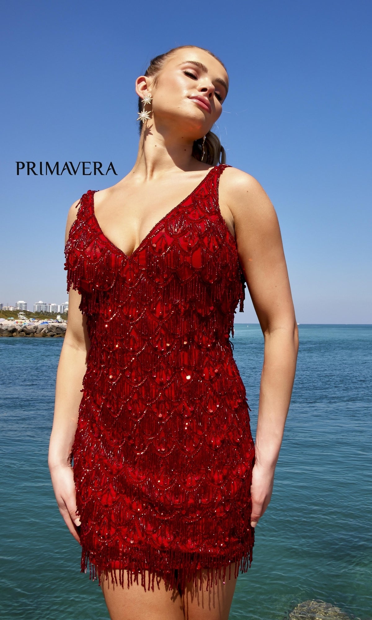 A woman in the Beaded-Fringe Short Semi-Formal Dress 4042 stands by the water, cityscape behind her under a clear blue sky. The word